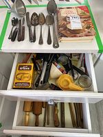 Photo showing the open drawer containing wooden rolling pins, plastic and glass measuring cups, metal funnel, and various other kitchen tools.