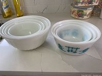 Four nested Fire King ovenware mixing bowls, three plain white and one with blue kitchen utensil design.