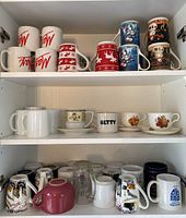Three shelves filled with various mugs including Christmas-themed, plain white mugs, printed mugs, and cup/saucer sets