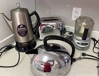 Photo showing all four small kitchen appliances together on a counter: Cuisinart cordless percolator, GE metallic plug-in toaster, Cuisinart food processor, and GE stainless steel kettle.