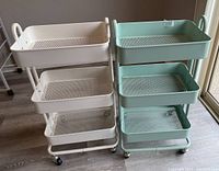Two metal mesh storage trolleys side by side on a wood floor near a wall and glass door. Shows overall size, structure and colors of the trolleys.