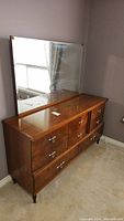 Photo of front view of wooden dresser with mirror reflecting window and part of room