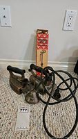 Two vintage irons, a small bell, a box with a brass toilet part, and an auction tag on a carpeted floor.