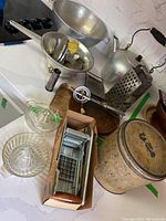 Photo showing assortment of vintage kitchen items including aluminum kettle, metal strainer, grater, potato masher, ceramic crock, and boxed potato cutter on a countertop.