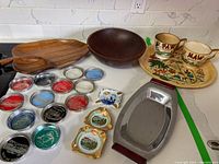 Wooden serving bowls and platters, souvenir coasters, ceramic ring holders, tropical-themed mugs, and a silver metal tray arranged on a white counter