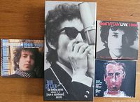 Four Bob Dylan Bootleg Series box sets arranged on a wooden surface showing different cover arts including a large black and white photo of Dylan with a harmonica, a portrait, and painted artwork.
