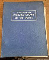 Cover of the blue textured album titled 'The Ambassador Album Postage Stamps of the World' showing wear consistent with age