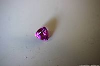 Close-up image of a small trillion cut red beryl gemstone lying on a plain surface, showing a deep pink-red color and faceted surface.