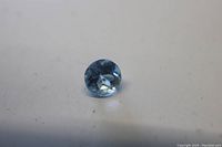 Close-up photo of a small round faceted aquamarine gemstone showing its light blue color and clarity on a white background.