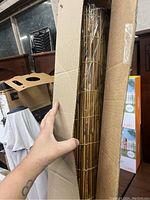 Photo of the bamboo privacy screen rolled up in cardboard packaging, showing natural bamboo slats and a hand holding the roll for scale.