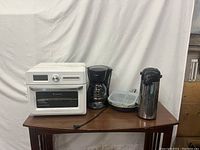 Full lot showing white multi-function appliance, black coffee maker, stainless steel thermos, and food containers on wooden table
