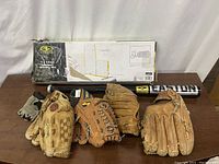 Baseball bat, four gloves, and boxed pitching net arranged on a wooden surface under white backdrop.