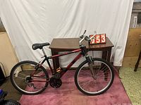 Side profile of black and red Schwinn mountain bike showing frame, tires, seat, and handlebars in front of brown wooden table and white backdrop.