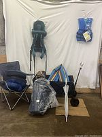 Photo of camping lot including folded tent in bag, camping chair with sitting pads, baby carrier, life jacket, wagon box, umbrella, and striped mat