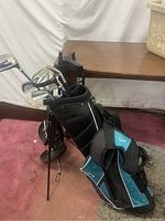 Golf bag standing on carpet with several golf clubs inside, showing black and teal color with stand extended.