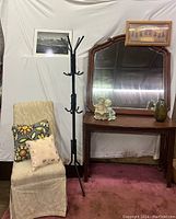 Photo showing a beige fabric-covered chair with two decorative pillows, a black metal coat rack, a dark wooden console table, large framed mirror, angel figurine, green glass vase, and a framed art print above the mirror.