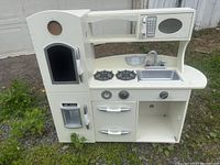 Full front view of the white wooden kids play kitchen showing refrigerator, microwave, stovetop, sink, oven, drawers, and open storage.