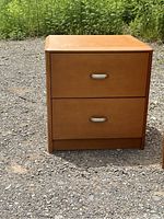 Front view of a light brown wooden night stand with two drawers and silver handles placed outdoors on gravel ground