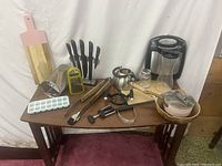 Full view of lot showing waffle maker, ice tray with cover, cutting boards, knives in block, kettle, cheese grater, tongs, potato masher, pan with lid and other kitchen utensils on a table.