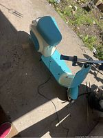 Photo of the blue and white Razer Vespa electric scooter on a concrete floor with greenery nearby, showing the entire scooter from an angled side top view.