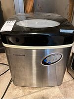 Front view of compact icemaker with brushed stainless steel body and black top. Shows control panel and non-detailed surroundings.