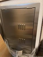 Front view of the stainless steel horizontal metal door showing ventilation slots and hinge on top.