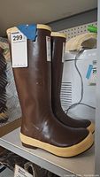 Pair of brown XtraTuf women's rubber boots with beige sole and trim, standing upright on a shelf with price tag visible.