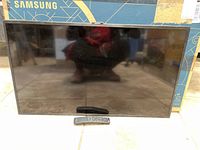 Front view of Samsung 32-inch Smart TV with remote control placed in front and original box in background.