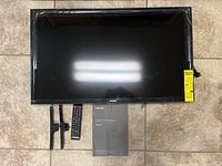 Samsung 32-inch TV laid flat with remote, two plastic TV stands, and user manual visible on tiled floor.