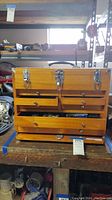 Front view of wooden tool chest showing multiple drawers and metal clasps, with some drawers open revealing tools and hardware.