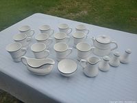 Full set of 20 white fine china pieces including tea cups, teapot with lid, creamers, sugar bowl, sauce boat, bowl, and salt and pepper set arranged on a table outdoors.