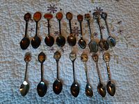 Image of 15 assorted collector spoons arranged in two rows on a quilted floral fabric. Each spoon has a unique emblem or design on the handle top.
