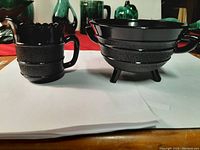 Pair of antique black pressed glass pieces on a table: milk jug with handle and ruffled spout on the left, sugar bowl with two handles and tripod legs on the right. Both have textured bands and glossy finish.