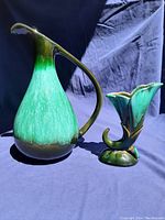 Two blue-green glazed pottery items on dark blue background showing a large marked jug with curved handle and smaller ruffled vase.