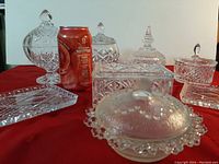 Four crystal lidded candy dishes of varying shapes and sizes arranged with a soda can for scale; includes round and oval shapes with finial lids on red cloth.