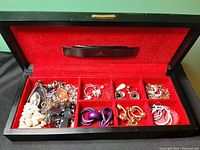 Top view of the open black jewelry box with red interior showing compartments filled with earrings, bracelets, and necklaces.