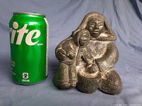 Side view of the full soapstone carving beside a soda can for scale, showing the seated mother and child with detailed texture on clothing and basket.