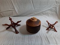 Two hand carved wooden trivets and one turned wooden bowl with lid, arranged on white fabric background.