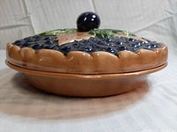 Side view of the pie plate with the lid on showing the berry and leaf design in dark blue and green glazes on a beige background, and the knob handle on the lid.
