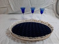 Overall view of the round vintage metal tray with blue crushed velvet lining and three blue color gradient aperitif glasses placed on a white cloth.