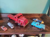 Full view of five vintage metal toy vehicles arranged on wooden surface. Includes a red flatbed truck, an orange race car, grey tanker truck, blue Corvette car, and a clown figure toy.