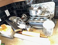 Wide view showing various metal baking pans, muffin tins, and trays stacked together with rolling pins and a plastic container with a blue lid on the counter.