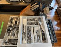 Multiple white plastic trays filled with various stainless steel forks, knives, spoons, and serving utensils, plus a butcher block with knives.