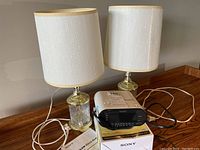 Two matching table lamps with white shades and metal glass bases alongside a Sony Dream Machine clock radio placed on a wooden surface.
