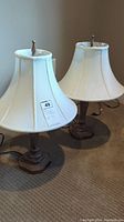Pair of table lamps with white fabric shades and brown bases, standing on carpet.