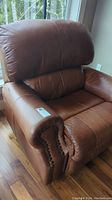 Front-left side view of brown leather recliner chair showing cushioned seat, padded backrest, and nailhead trim on arm.