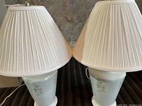 Full view of two matching ceramic base table lamps with floral design and pleated fabric lampshades on dark carpet.