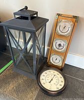 Photo of large black metal hurricane lantern with cross-braced glass panels and a wooden weather instrument panel with three brass-finished gauges.