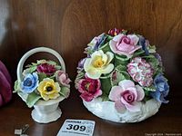 Photo of two fine china flower baskets by Aynsley, one with handle and one larger basket with varied flowers, displayed on wooden shelf.