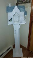 Front view of white wooden birdhouse with blue roof on pedestal stand indoors.
